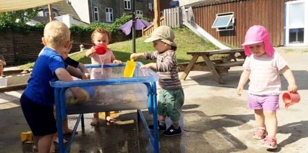 Børn leger med vand på vuggestuens legeplads en varm sommerdag 