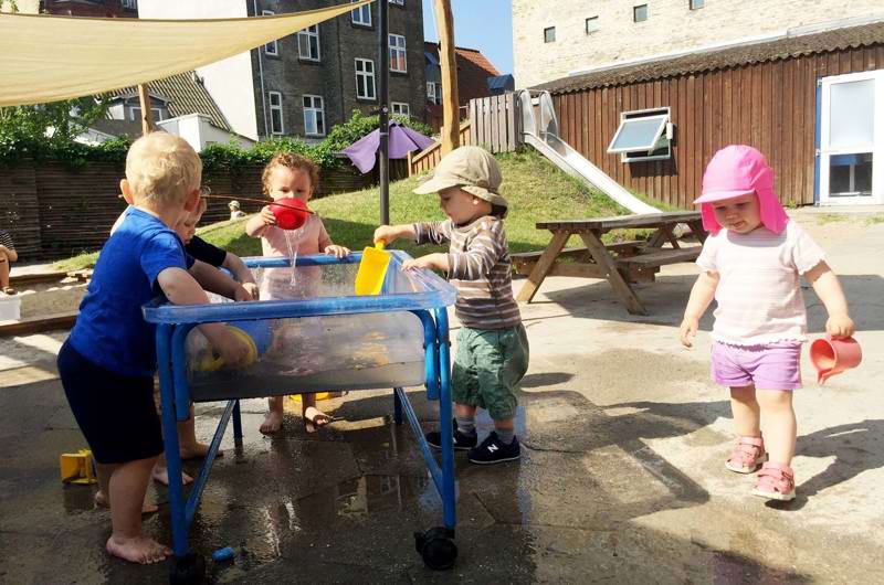 Børn leger med vand på vuggestuens legeplads en varm sommerdag